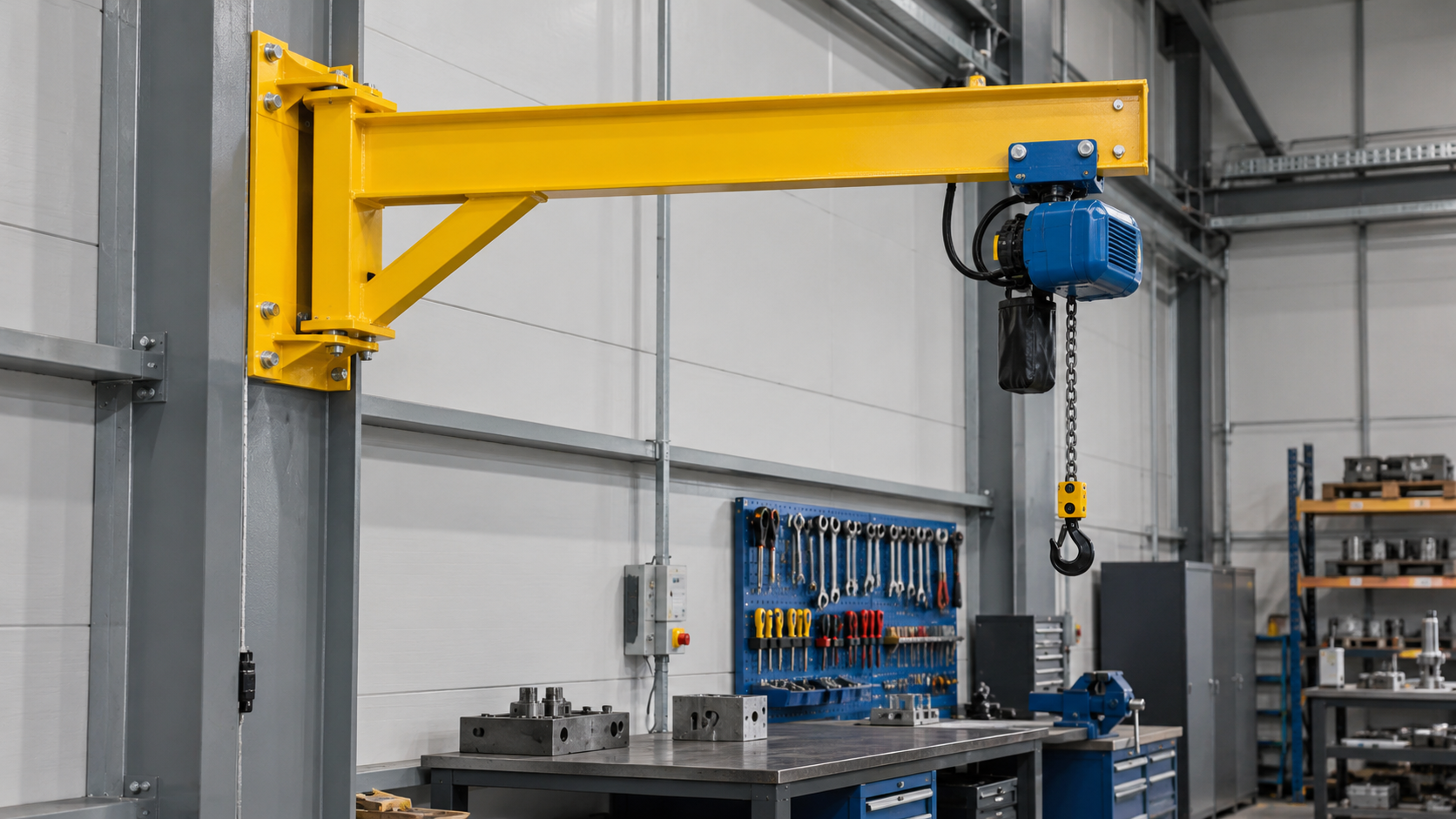 One yellow wall mounted jib crane with a short horizontal arm and blue electric chain hoist at a compact fabrication workstation, black hook unloaded, fixed grey steel wall column and a tidy assembly bench below.